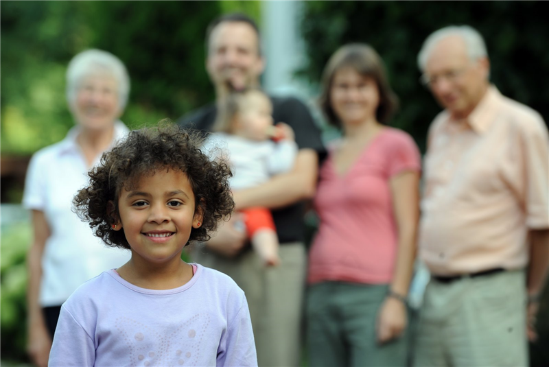 dunkelhäutiges Kind steht im Vordergrund, im Hintergrund ist eine hellhäutige Familie zu erkennen