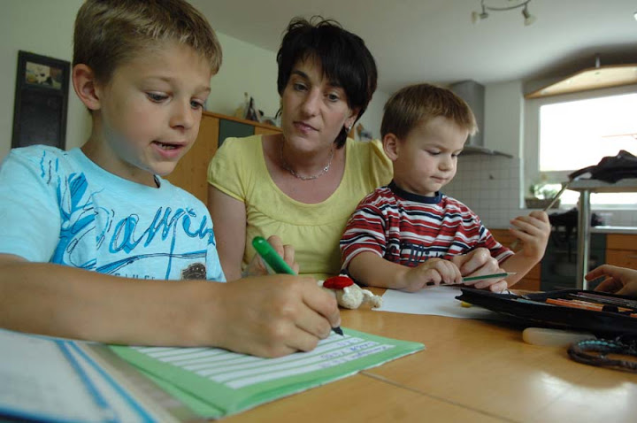 Eine Frau schaut ihrem Sohn bei den Hausaufgaben zu, ein kleineres Kind sitzt auf ihrem Schoß