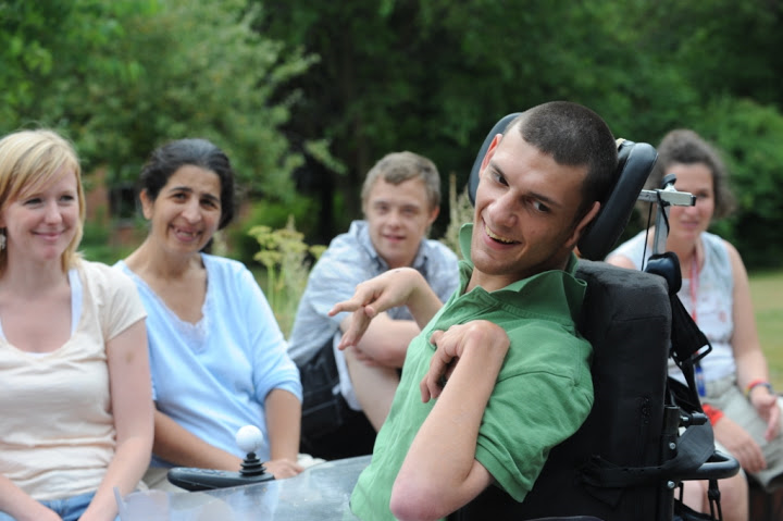 Ein Mann mit Behinderung im Vordergrund, im Hintergrund sitzt eine Gruppe Menschen